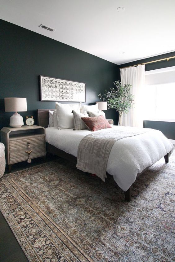 a boho master bedroom with dark green walls, a cool bed and eye-catchy nightstands, a potted plant and printed textiles