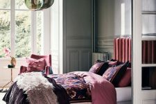 a bold refined bedroom with grey molding walls, a pink upholstered bed and a pink chair, bright floral bedding and a dreamy pendant lamp