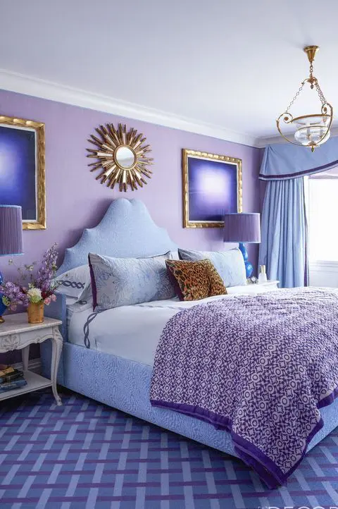 a bright purple bedroom with a lilac accent wall, gold details, a pretty pendant lamp, some refined nightstands and a sunburst mirror