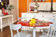 a colorful eat-in kitchen with an orange accent wall, white cabinets, a bright dining space with a round red table and chairs with colorful legs