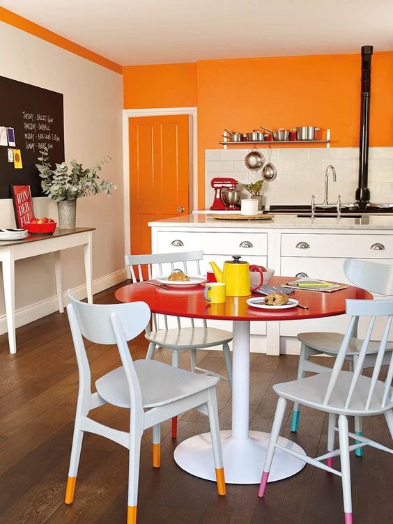 A colorful eat in kitchen with an orange accent wall, white cabinets, a bright dining space with a round red table and chairs with colorful legs