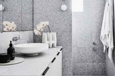 a contemporary bathroom clad with grey terrazzo tiles, with a white vanity, a marble countertop, a round sink, a cool vase and orchids