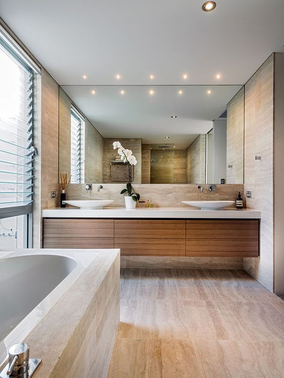 A contemporary bathroom clad with neutral stone imitating tiles, a floating vanity, a large mirror, a glazed wall with shades
