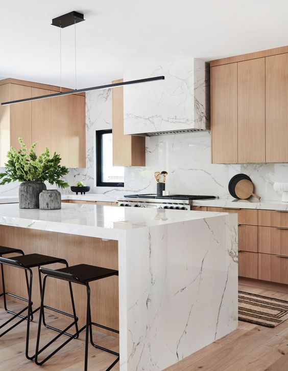 A contemporary kitchen with elegant light stained cabinets, a white stone hood and countertops plus a bakcsplash and a matching kitchen island