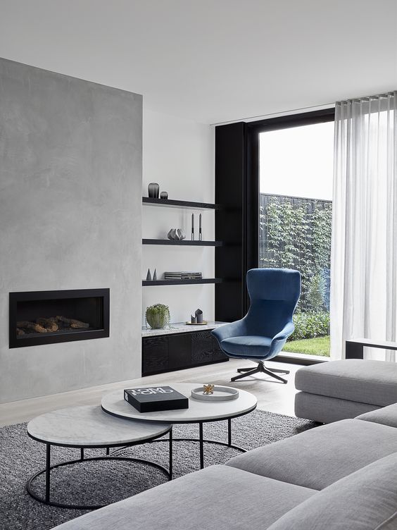 A contemporary living room with a concrete fireplace, built in shelves, a blue chair, a grey sectional and a duo of tables