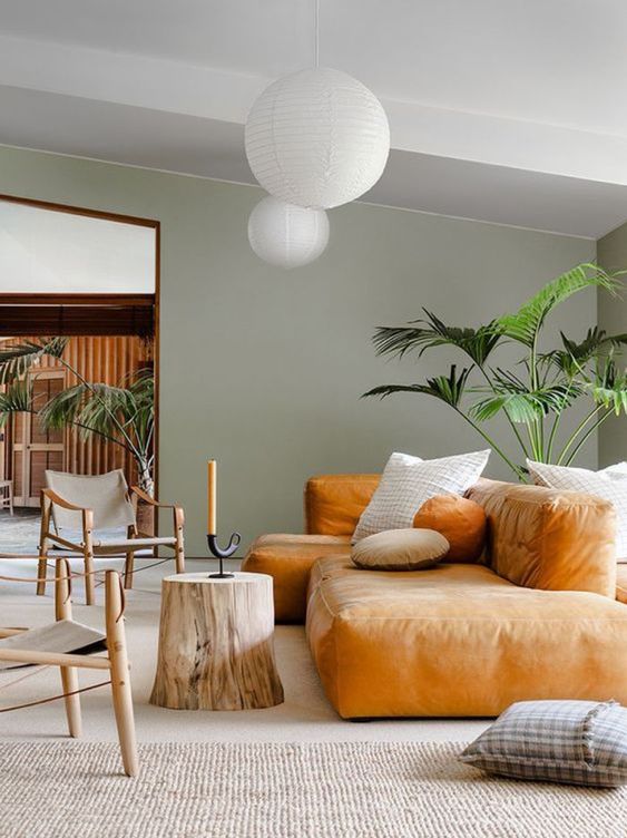 A contemporary living space with an amber colored low sofa, a tree sutmp, catchy wooden chairs, statement plants and pendant lamps