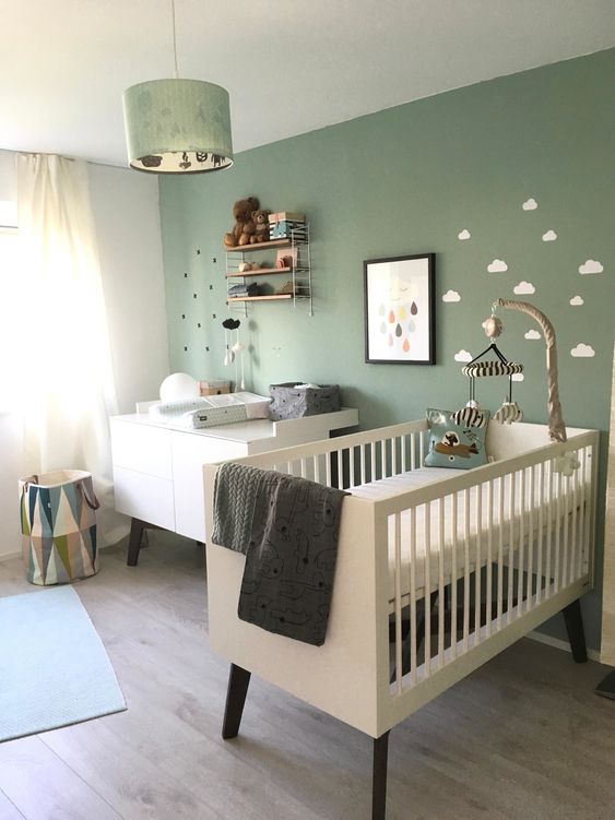 a cozy Nordic nursery with a sage green accent wall, white furniture, a pendant lamp and some shelves plus neutral bedding