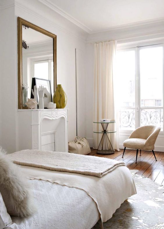a dreamy Parisian style bedroom with a non-working fireplace, a cool chair and a round table, a large mirror in a gilded frame and neutral textiles