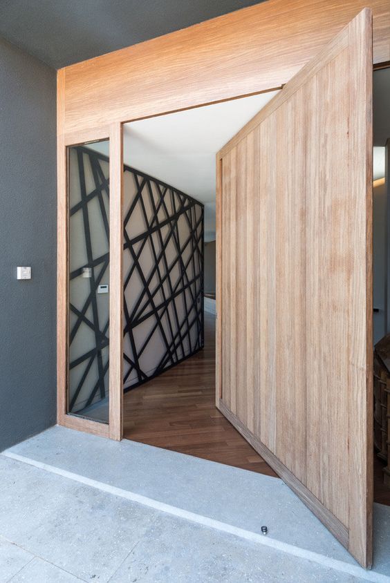 a large light-stained wood pivot door with glass panes and no handles or knobs is a very cool idea for a minimalist entrance