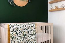 a little and lovely nursery with a dark green accent wall, a stained crib, open shelves and a cool decoration of wood and eucalyptus