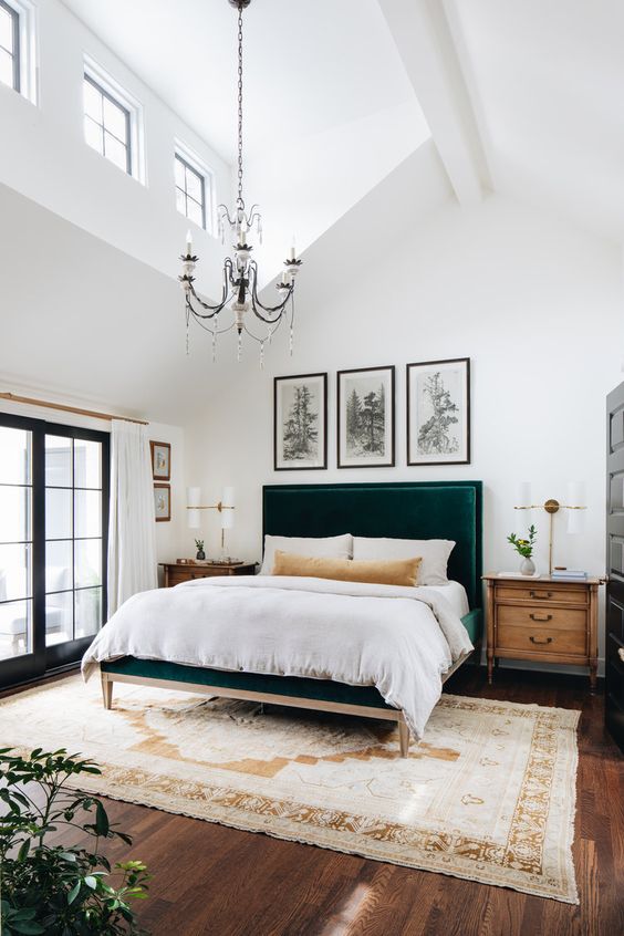 a lovely bedroom with a double height ceiling, a vintage chandelier that highlights it, a green upholstered bed, a grid gallery wal and stained nightstands