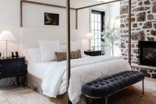 a lovely bedroom with a farmhouse feel, a stone clad fireplace, a canopy bed, an upholstered bench, black nightstands, neutral bedding and a potted tree