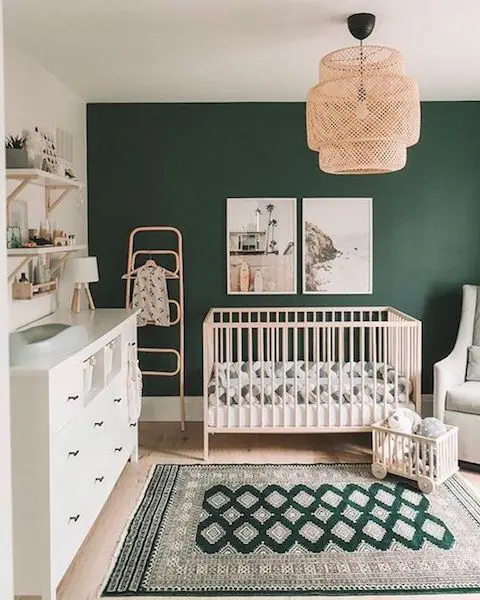 a lovely gender neutral nursery with a dark green accent wall, neutral furniture, a woven pendant lamp, some open shelves and printed bedding