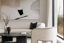 a luxurious contemporary living room in neutrals and black, with a curved sofa and a chair on three legs, a black marble table, a chandelier plus a statement artwork