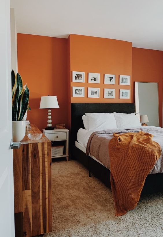 A mid century modern bedroom with an orange accent wall, a black bed and neutral nightstands, a reclaimed wood dresser and a small gallery wall