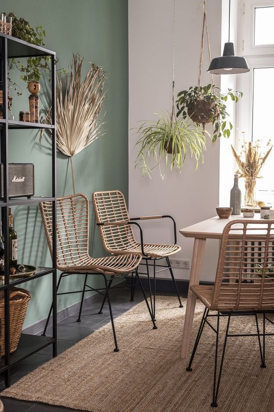 a modern boho dining space with a green accent wall, a light stained table and rattan chairs, a black shelvint unit and potted greenery