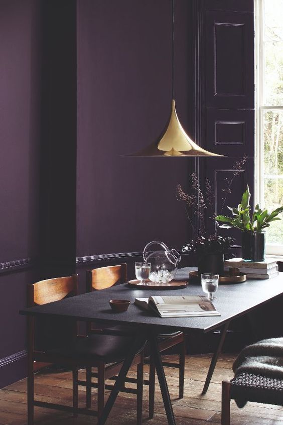 a moody dining room with a deep purple accent wall, a black trestle table and wooden chairs, a shiny gilded pendant lamp