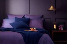 a moody monochromatic bedroom with a deep purple paneled wall, purple and navy bedding, a wooden nightstand and a metal pendant lamp