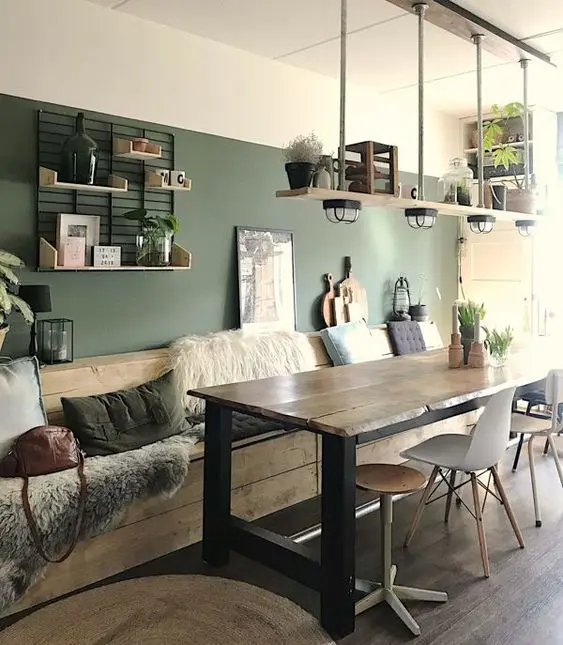 A rustic dining room with a green accent wall, a wooden table and mismatching chairs, a wooden built in bench with lots of pillows