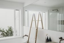 a serene white contemporary bathroom with an oval tub, a floating vanity, a long and large mirror and a ladder with towels