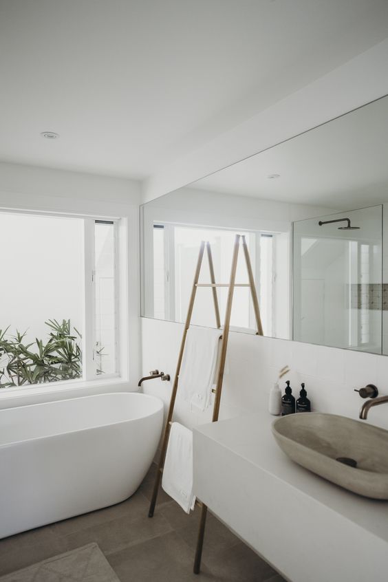 a serene white contemporary bathroom with an oval tub, a floating vanity, a long and large mirror and a ladder with towels