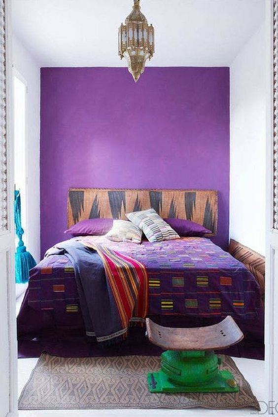 a small and colorful boho bedroom with a purple accent wall, a bed with bright bedding, a small stool and a Moroccan pendant lamp