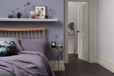 a stylish modern bedroom with a lilac accent wall, a wooden bed with lilac and grey bedding, a floating shelf with lovely decor