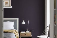 a stylish moody bedroom with a dark purple accent wall, grey and white furniture, a small nightstand with a table lamp and ledges with artworks