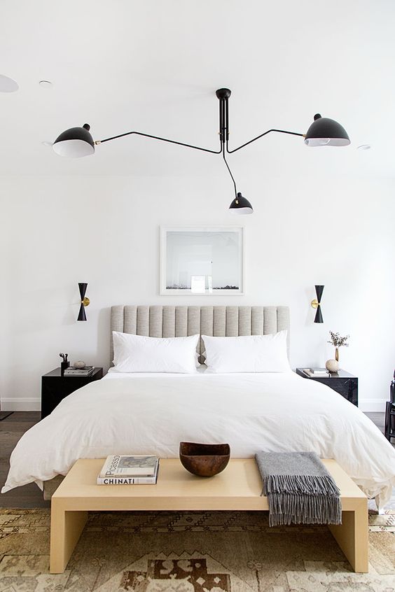 a stylish neutral bedroom with a grey upholstered bed, a wooden bench, dark matching nightstands and a black chandelier