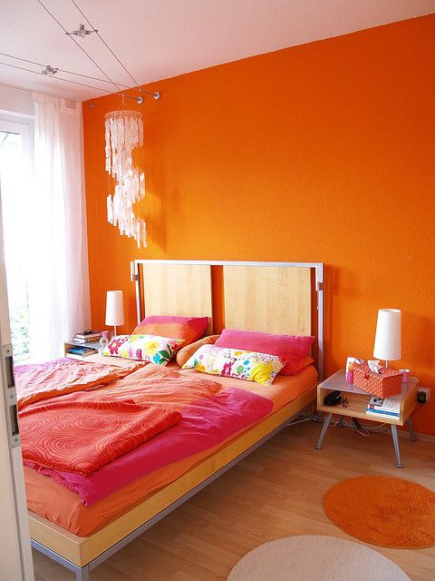 a super colorful bedroom with an orange accent wall, chic mid-century modern furniture, bold bedding, a crystal chandelier and bold rugs