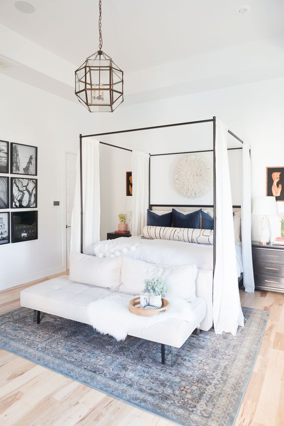 a welcoming bedroom in neutrals with a canopy bed and curtains, a grid gallery wall, black nightstands, an upholstered bench and pillows