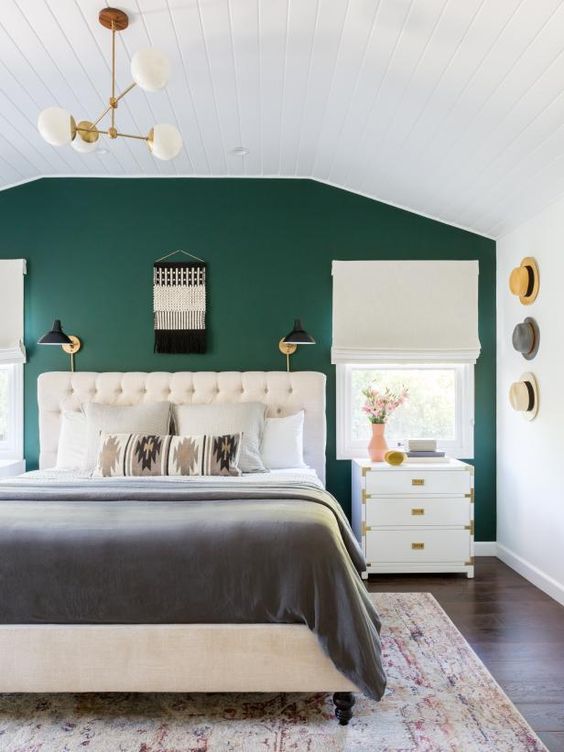 a welcoming boho bedroom with an emerald accent wall, a creamy upholstered bed, white nightstands, a macrame decoration and hats as decor