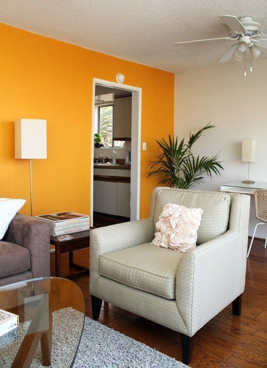 an elegant modern living room with a bright orange accent wall, a grey sofa and a neutral printed chair, a cool desk and a woven chair plus a glass table