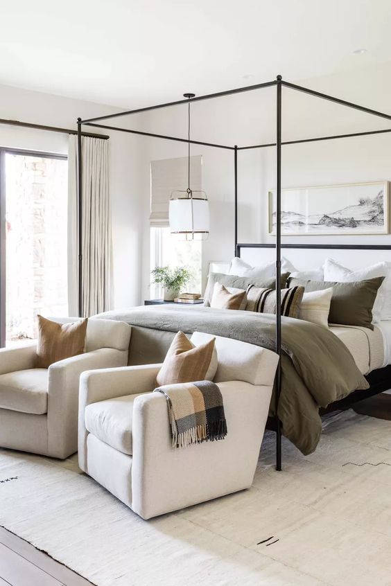 an inviting neutral modern bedroom with a canopy bed, muted and printed bedding, a pendant lamp, creamy chairs  and an artwork