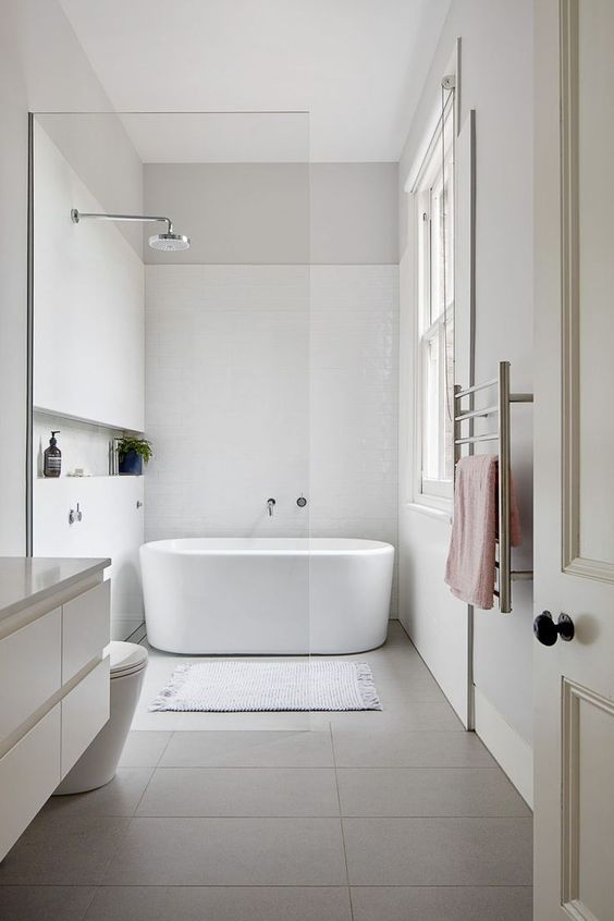 An ultra minimalist bathroom with white and grey tiles, an oval tub, chrome fixtures and pastel textiles and a window for more light