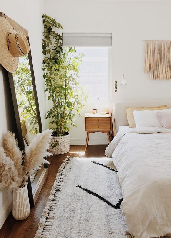 A beautiful and light filled bedroom with an upholstered bed, a floor mirror, macrame hanging, pampas grass and a potted plant