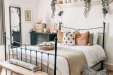 07 a stylish boho bedroom with a metal bed, a black dresser, a wooden bench, potted greenery, a woven lamp, a basket and a floor mirror