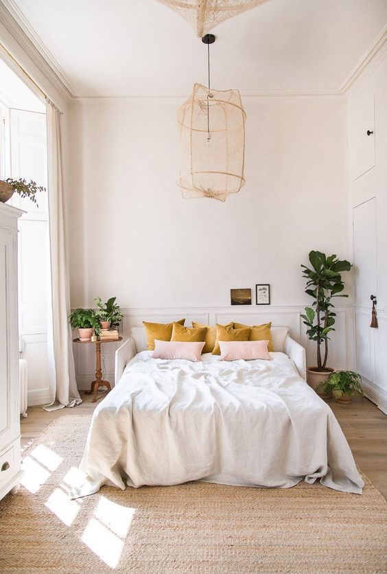 A pretty light filled bedroom with paneled walls, a white bed, a pendant lamp, mustard and pink pillows and some potted greenery to refresh the space