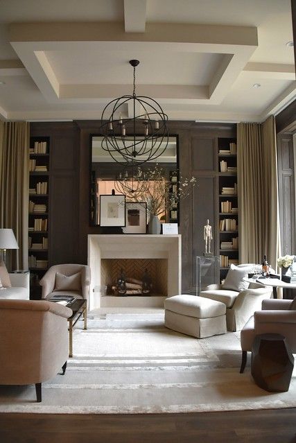 A sophisticated taupe living room with paneling, built in bookshelves, a non working fireplace, neutral seating furniture, a sphere pendant lamp and a large mirror