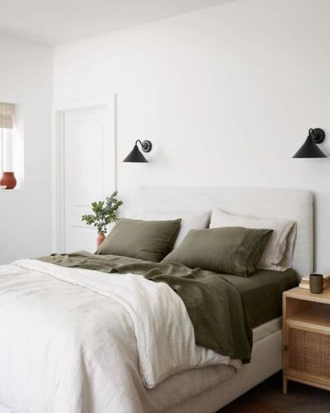 a pretty bedroom with a neutral upholstered bed, green and neutral bedding, nightstands, black sconces and potted greenery