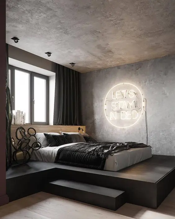 a contemporary industrial bedroom with a bed on a platform, black and white bedding, a neon sign, dark curtains and concrete walls and a ceiling