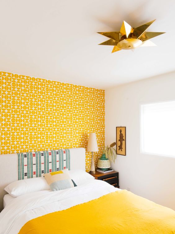 a bright bedroom with a yellow printed wallpaper wall, a neutral bed with a statement headboard and a yellow blanket, a pretty artwork is chic