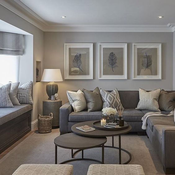 a cozy small grey living room with a windowsill daybed with printed pillows, a taupe sectional, a duo of brown round tables and a cool gallery wlal