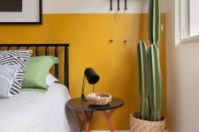 20 a modern boho bedroom with color block yellow and white walls, a blakc metal bed, a round table, a potted cactus and some art and pillows