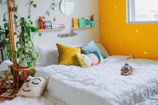 21 a pretty bedroom with a yellow accent wall, a low bed on the floor, climbing up plants, a macrame hanging, shelves and colorful pillows