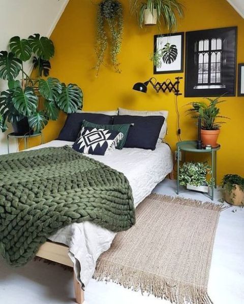 A pretty bedroom spruced up with Gen Z yellow   an accent wall, a bed with black, green and neutral bedding, potted plants, artworks and a green table is super cool