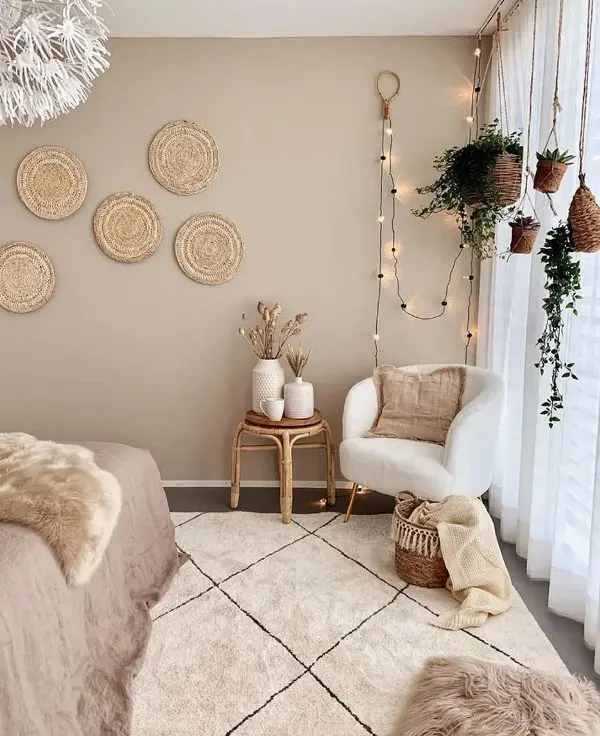 a boho bedroom with taupe walls, a bed, a creamy chair, a rattan stool, hanging potted greenery, decorative woven plates and layered rugs