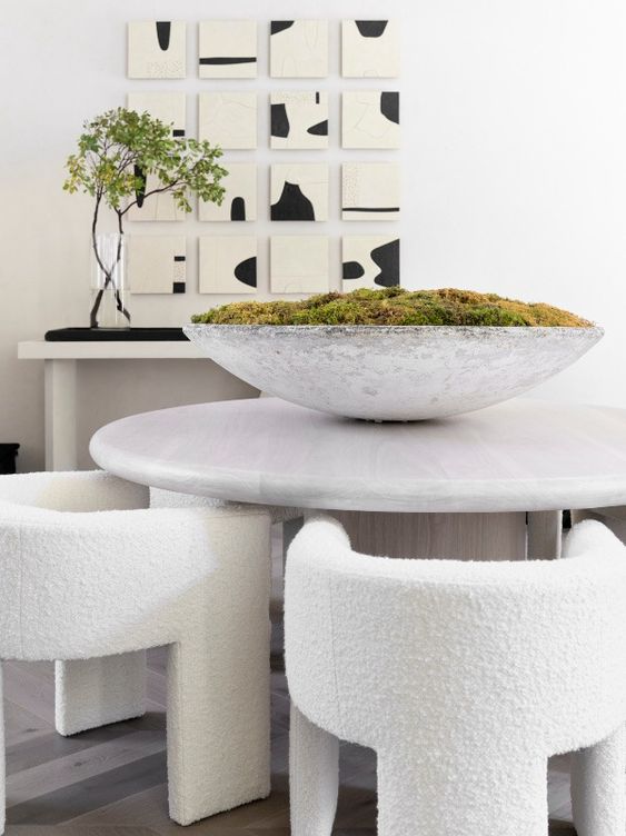a bold contemporary dining room with a neutral console and a catchy artwork, a round stone table, creamy rounded chairs and a bowl with moss