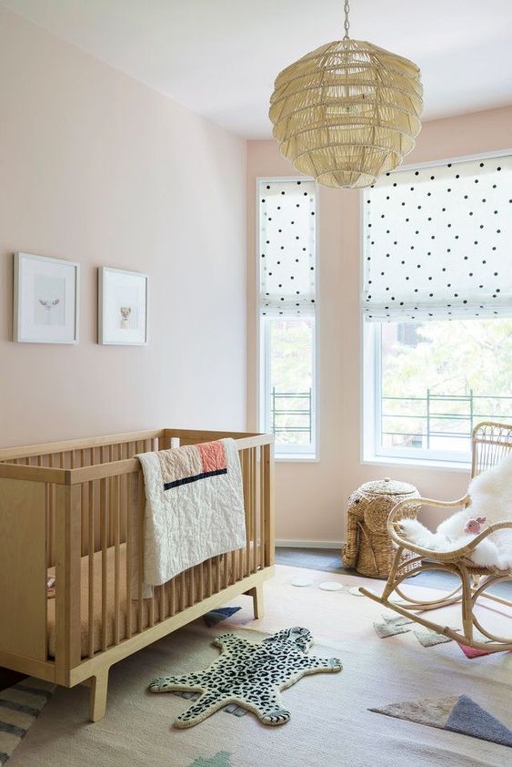 a catchy contemporary nursery with blush walls, stained wood and rattan furniture, a pendant lamp, layered rugs and polka dot shades