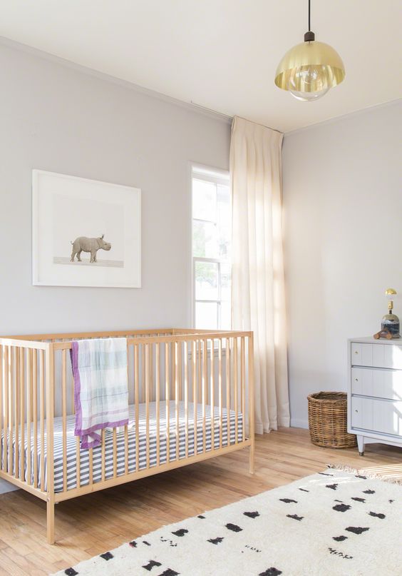 a chic and serene nursery with dove grey walls, a light-stained crib, a grey dresser, a printed rug and blanket, a gold pendant lamp